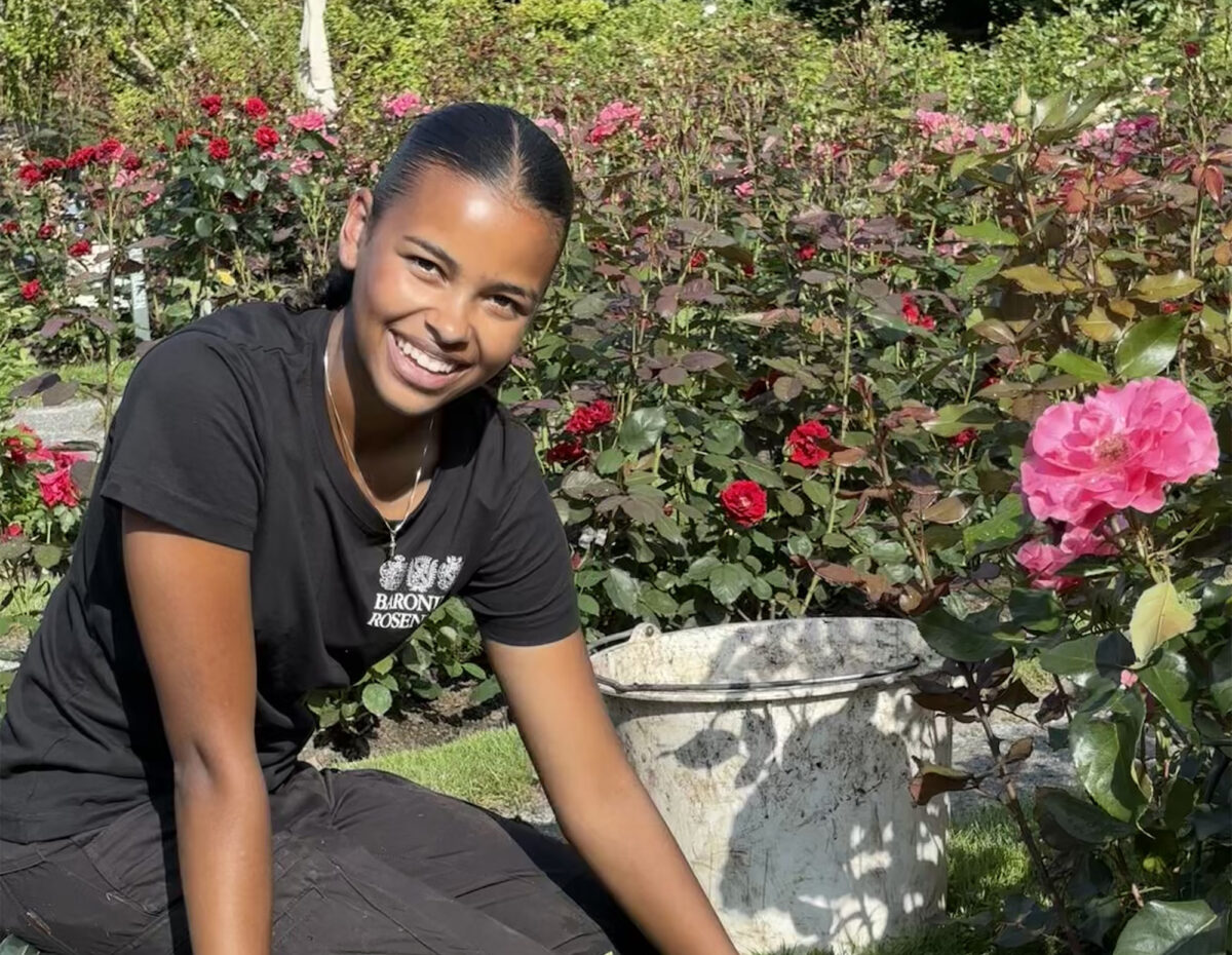 Ei jente med mørkt hår og klede sit på kne i ein hage framfor ei rose. Ho har hanskar på seg og smiler.