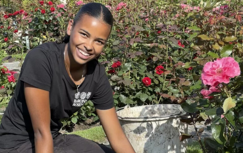 Ei jente med mørkt hår og klede sit på kne i ein hage framfor ei rose. Ho har hanskar på seg og smiler.