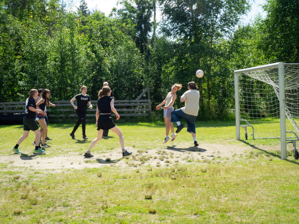 Fleire ungdomar speler fotball. Ei av dei heng i lufta, og ballen flyg mot mål.