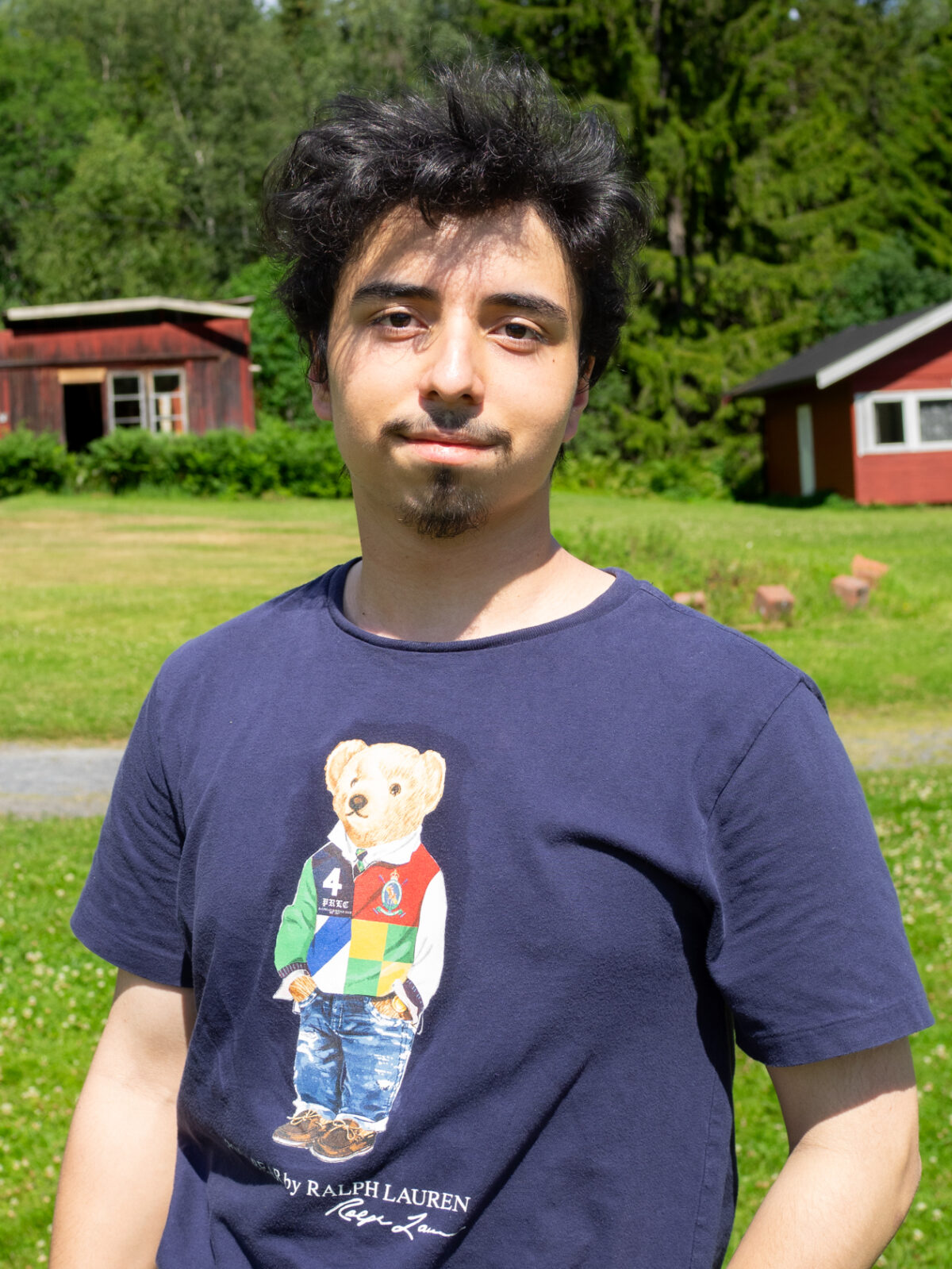 Emir Katan har mørkt hår og fippskjegg. Han har ein blå t-skjorte frå Ralph Lauren. Den har ein teddybjørn på.