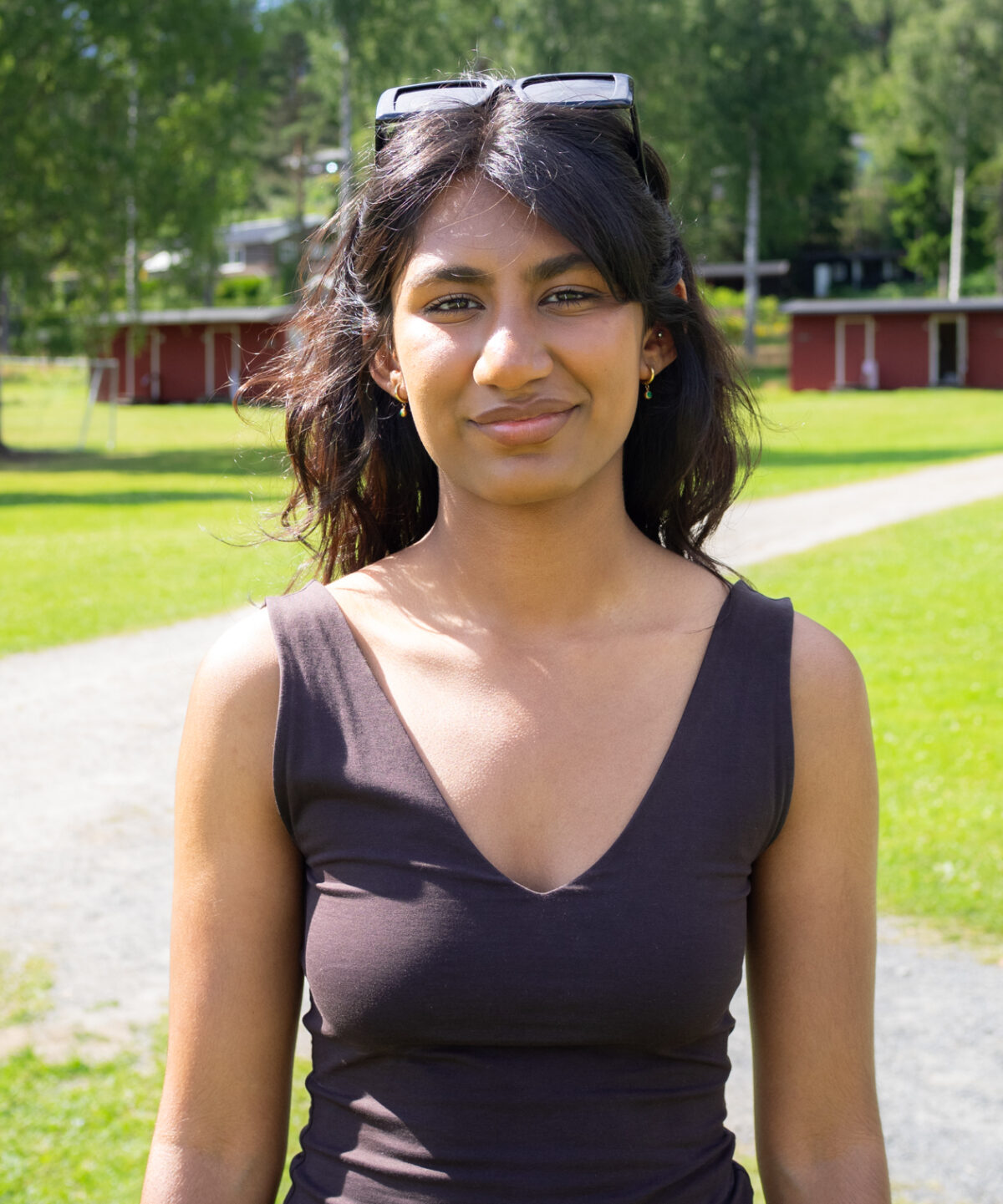 Sanjita har indisk bakgrunn. Ho står på ein plen i ein mørk topp med v-hals. Ho har brunt hår. Ho har eit par solbriller på hovudet.