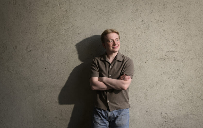Gut med lyst hår står med armene i kors og lener seg mot ein betongvegg. Han ser til sida med eit smil.